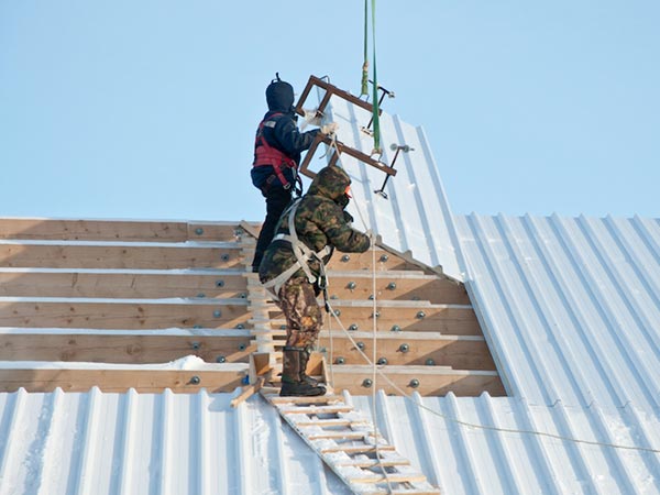 Two construction workers on a wooden roof frame, guiding a metal sheet into place with a lift and wearing safety gear.