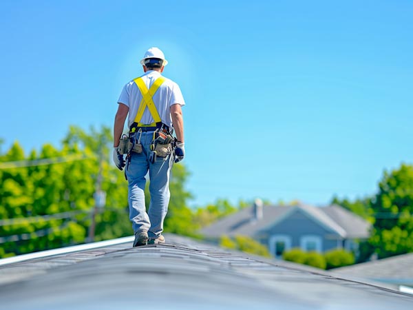 Construction worker in a white hard hat and yellow safety harness walking along a sloped rooftop with tools at his belt, residential neighborhood in the background.