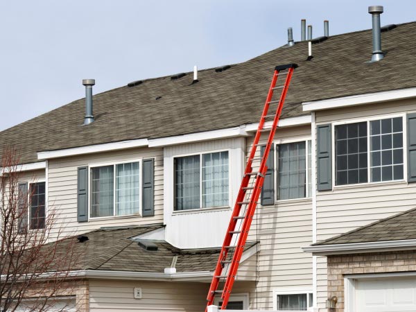 Red extension ladder leaning against a beige townhouse roof during maintenance, with vents visible on the roof.