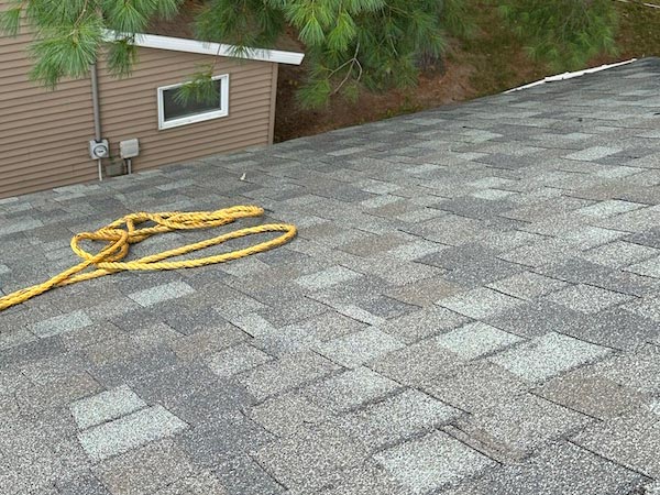 Coiled yellow rope lying on a gray-shingled roof near a house wall with a small window and overhanging evergreen branches above.